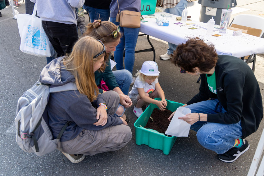 A child and three adults bending down to look into a bin containing topsoil and seeds
